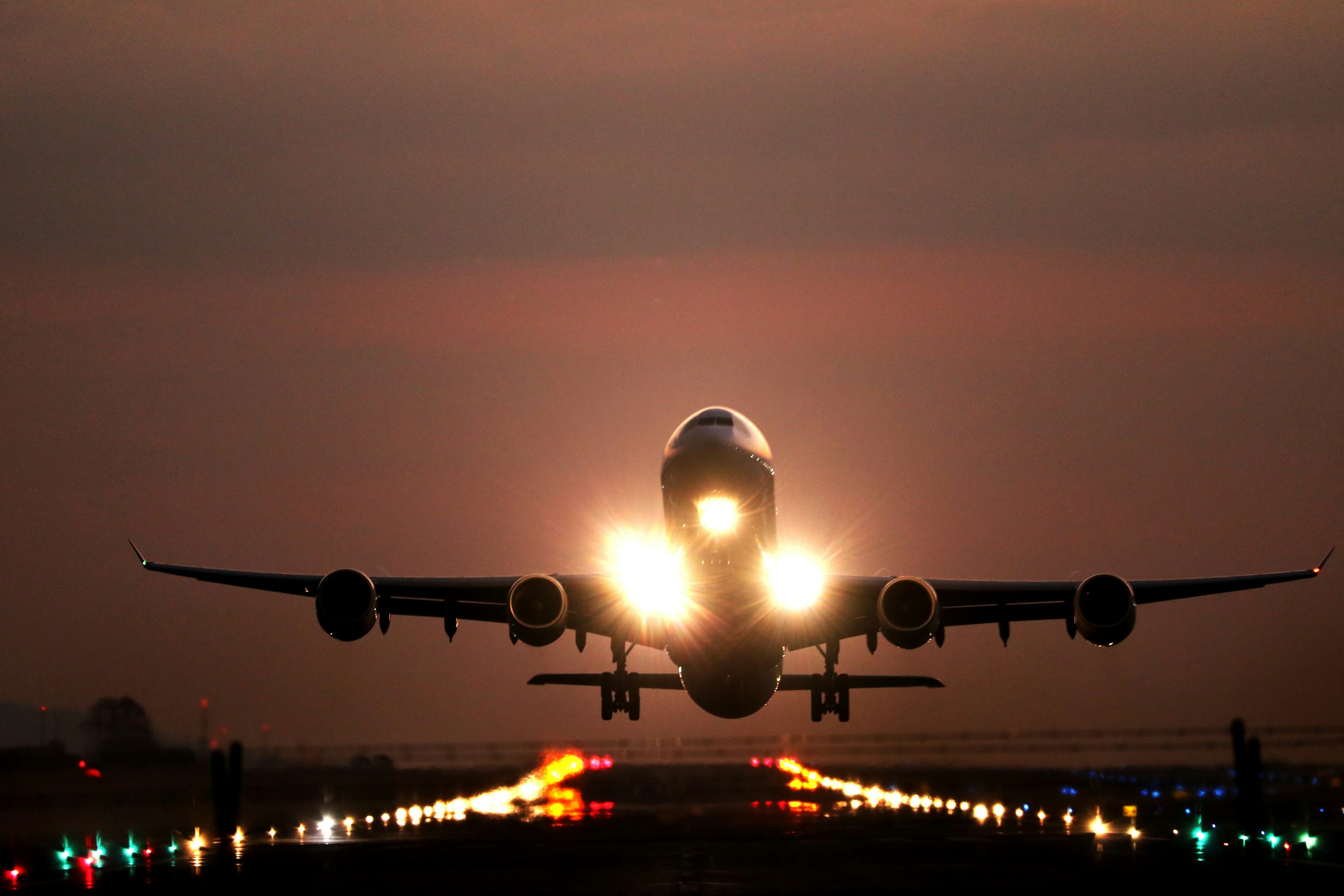 A four-engine wide-body airliner lifting off at dusk, landing lights blazing above the runway lights